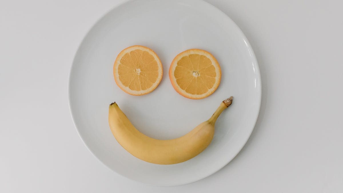 A playful smiley face made with a banana and orange slices on a ceramic plate, viewed from above.